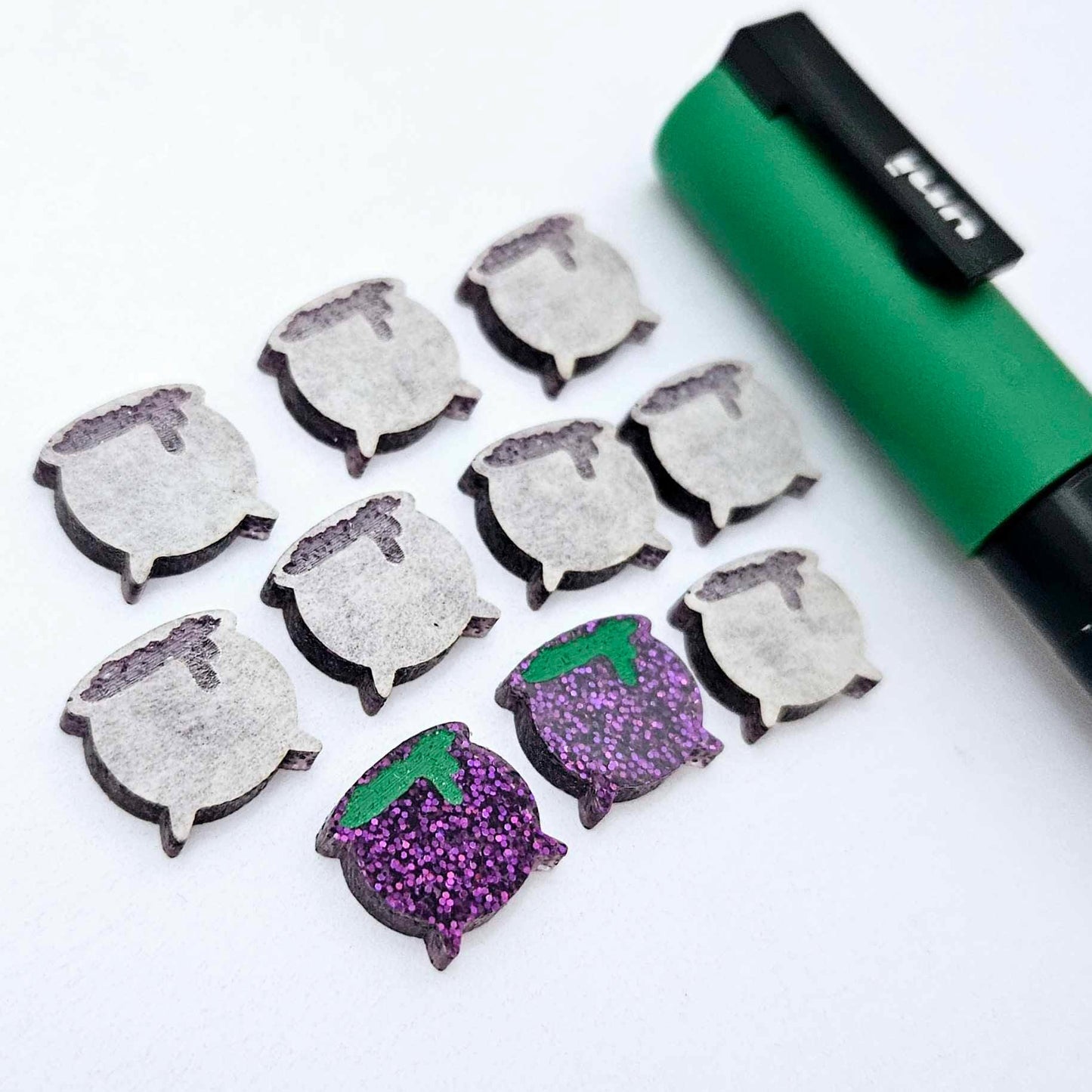 One pair of holographic purple glitter acrylic Witch's Cauldron stud earring blanks with green paint-filled details, surrounded by multiple unpainted pieces with protective paper on, pictured with a Green Posca Paint Pen