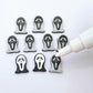 One pair of black glitter acrylic spooky face mask stud earring blanks with white paint-filled details, surrounded by multiple unpainted pieces with protective paper on, pictured with an uncapped White Posca Paint Pen ready for paint filling