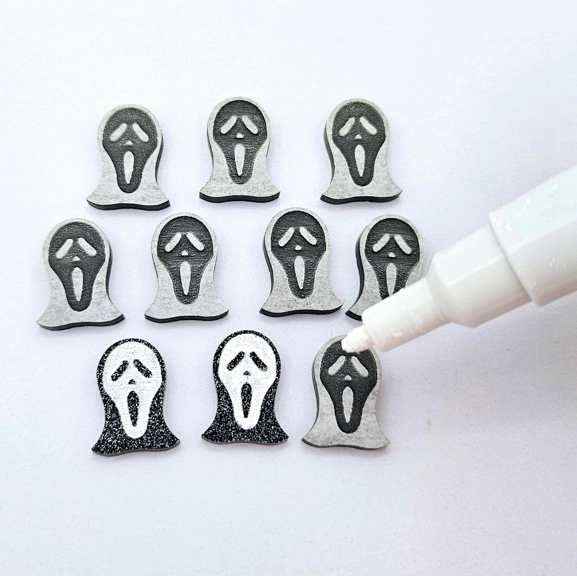 One pair of black glitter acrylic spooky face mask stud earring blanks with white paint-filled details, surrounded by multiple unpainted pieces with protective paper on, pictured with an uncapped White Posca Paint Pen ready for paint filling