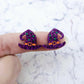 One pair of 18mm holographic purple glitter acrylic witch’s hat stud earring blanks with orange and green paint-filled details shown on a finger for size reference
