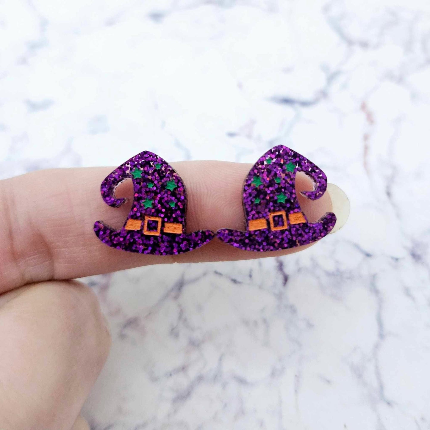 One pair of 18mm holographic purple glitter acrylic witch’s hat stud earring blanks with orange and green paint-filled details shown on a finger for size reference