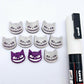 One pair of holographic purple glitter acrylic Spooky Cat stud earring blanks with white paint-filled details, surrounded by multiple unpainted pieces with protective paper on, pictured with a White Posca Paint Pen