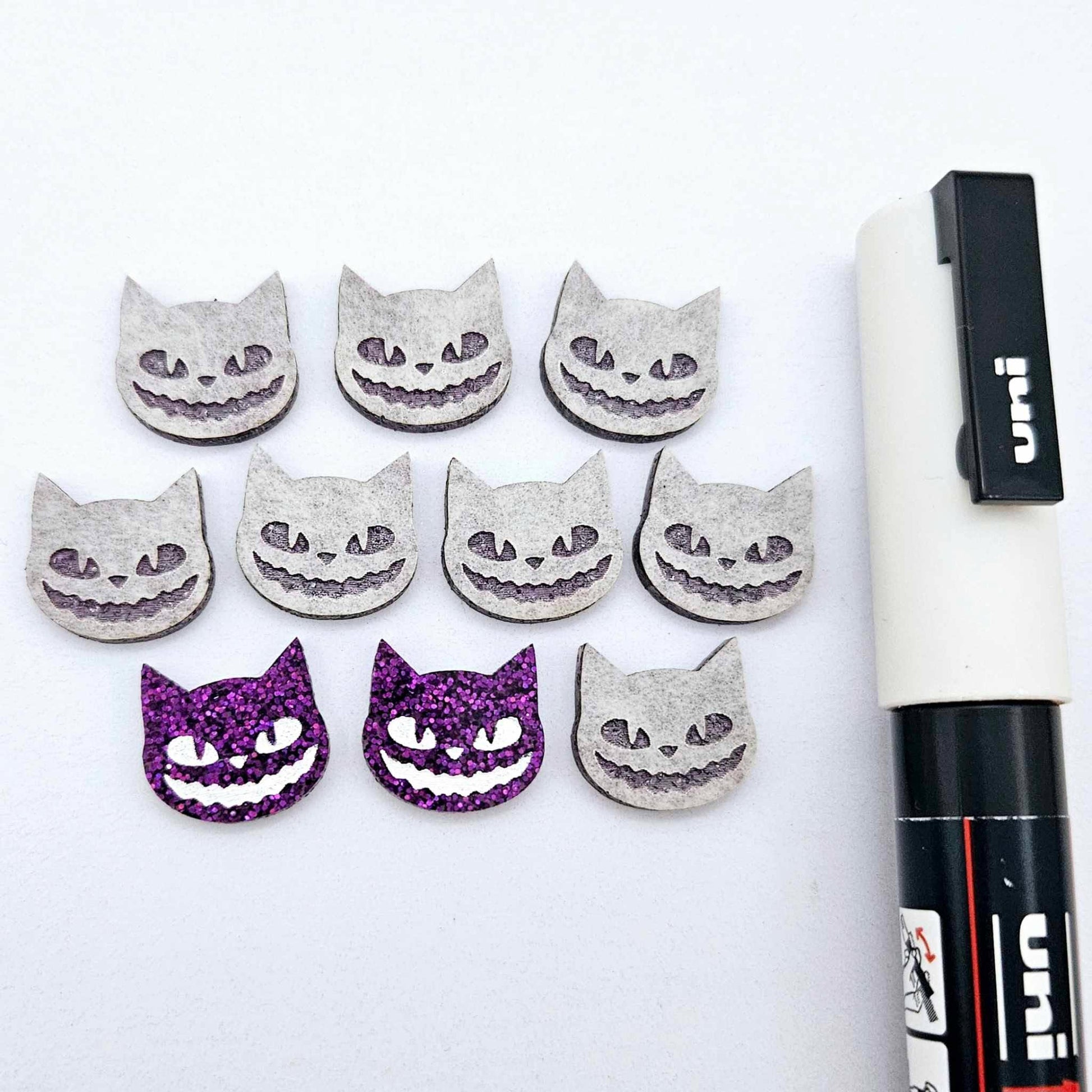 One pair of holographic purple glitter acrylic Spooky Cat stud earring blanks with white paint-filled details, surrounded by multiple unpainted pieces with protective paper on, pictured with a White Posca Paint Pen