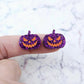 One pair of 16mm holographic purple glitter Jack-O’-Lantern stud earring blanks with orange paint-filled facial details shown on a finger for size reference