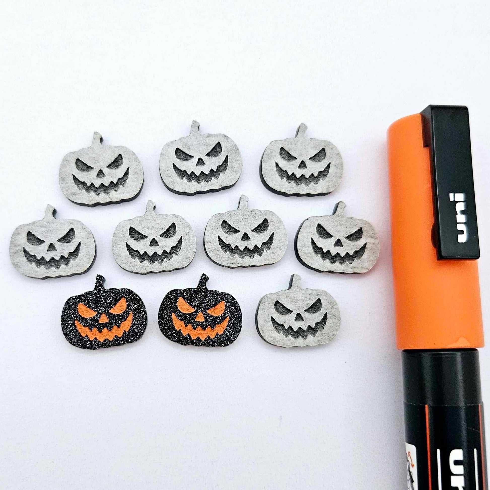 One pair of black glitter acrylic Jack-O’-Lantern stud earring blanks with orange paint-filled details, surrounded by multiple unpainted pieces with protective paper on, pictured with an Orange Posca Paint Pen