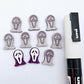One pair of holographic purple glitter acrylic spooky face mask stud earring blanks with white paint-filled details, surrounded by multiple unpainted pieces with protective paper on, pictured with a White Posca Paint Pen