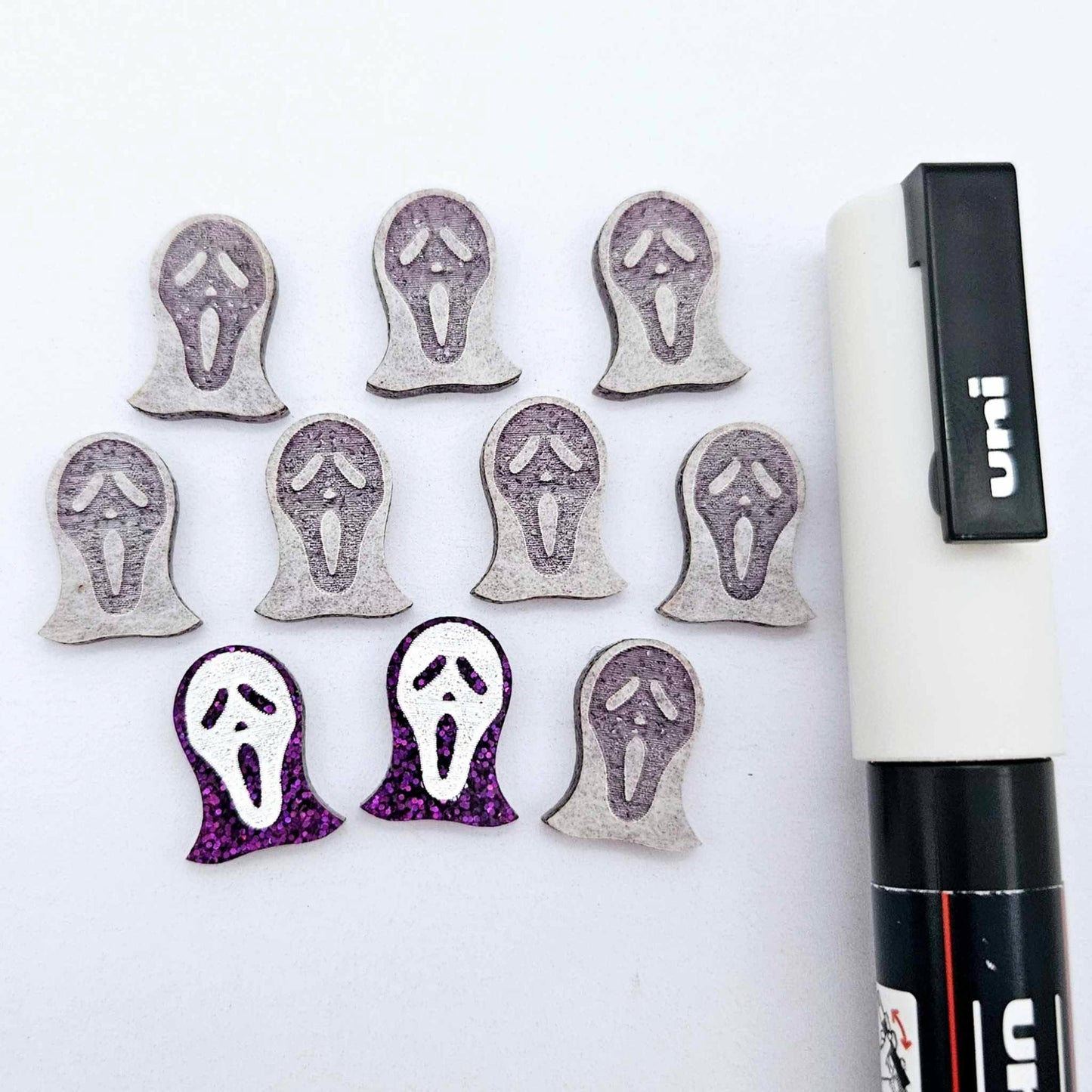 One pair of holographic purple glitter acrylic spooky face mask stud earring blanks with white paint-filled details, surrounded by multiple unpainted pieces with protective paper on, pictured with a White Posca Paint Pen