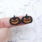 One pair of 16mm black glitter acrylic Jack-O’-Lantern stud earring blanks with orange paint-filled facial details shown on a finger for size reference