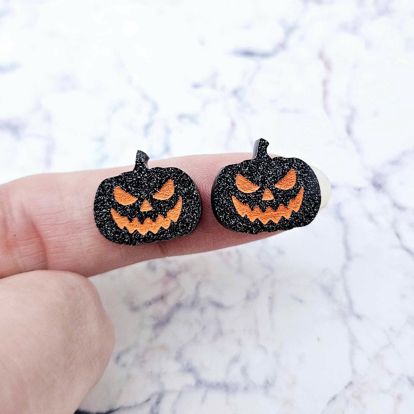 One pair of 16mm black glitter acrylic Jack-O’-Lantern stud earring blanks with orange paint-filled facial details shown on a finger for size reference