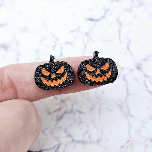 One pair of 16mm black glitter acrylic Jack-O’-Lantern stud earring blanks with orange paint-filled facial details shown on a finger for size reference