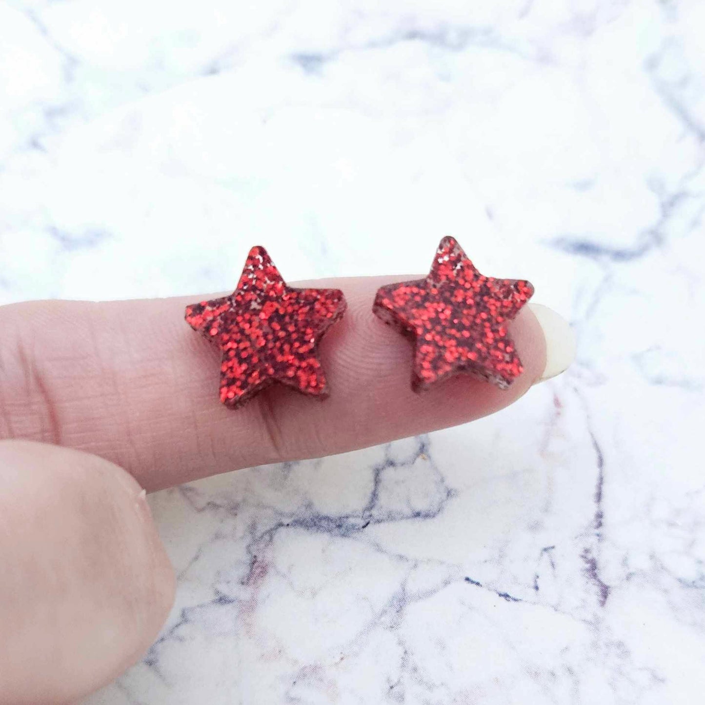 One pair of 14mm red glitter acrylic star stud earring blanks shown on a finger for size reference