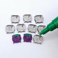 One pair of holographic purple glitter acrylic Witch's Cauldron stud earring blanks with green paint-filled details, surrounded by multiple unpainted pieces with protective paper on, pictured with an uncapped green Posca Paint Pen ready for paint filling