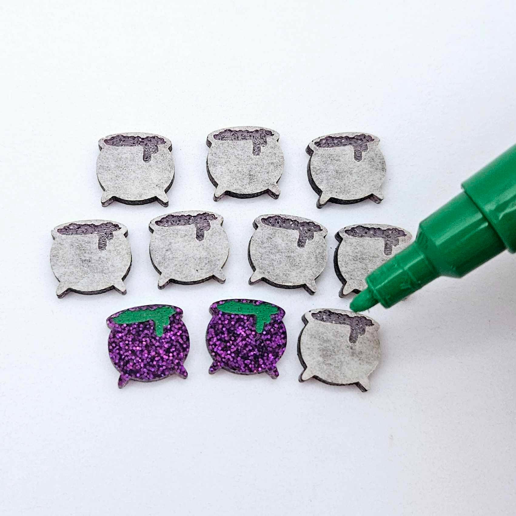 One pair of holographic purple glitter acrylic Witch's Cauldron stud earring blanks with green paint-filled details, surrounded by multiple unpainted pieces with protective paper on, pictured with an uncapped green Posca Paint Pen ready for paint filling