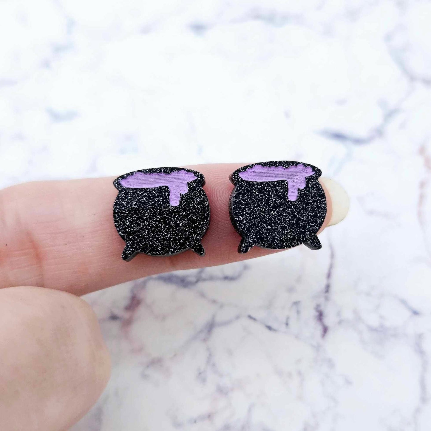 One pair of 14mm black glitter acrylic Witch's Cauldron stud earring blanks with lavender paint-filled bubbling potion details shown on a finger for size reference