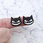 One pair of 16mm black glitter acrylic Spooky Cat stud earring blanks with white paint-filled facial details shown on a finger for size reference.