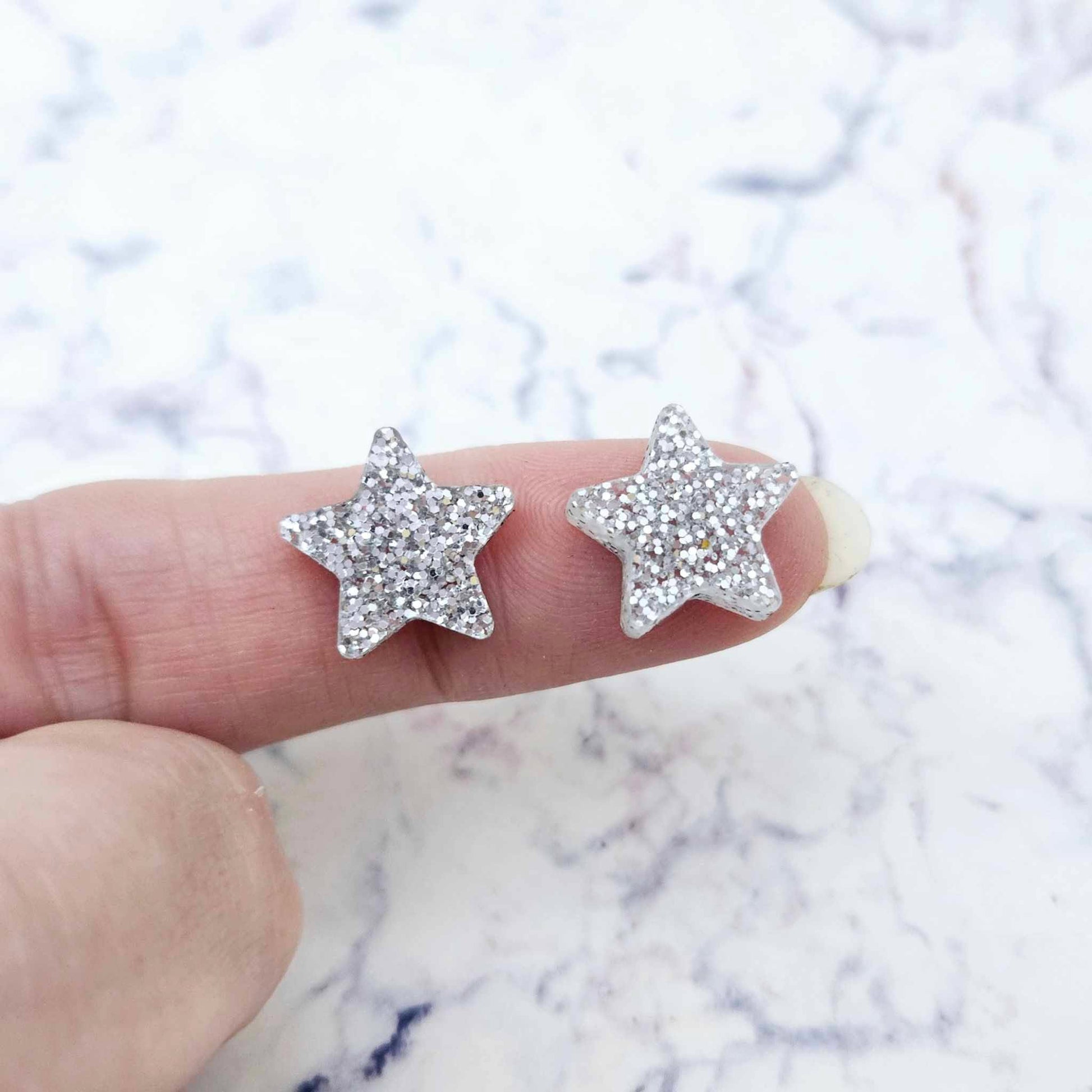 One pair of 14mm silver glitter acrylic star stud earring blanks shown on a finger for size reference