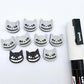 One pair of black glitter acrylic Spooky Cat stud earring blanks with white paint-filled details, surrounded by multiple unpainted pieces with protective paper on, pictured with a White Posca Paint Pen