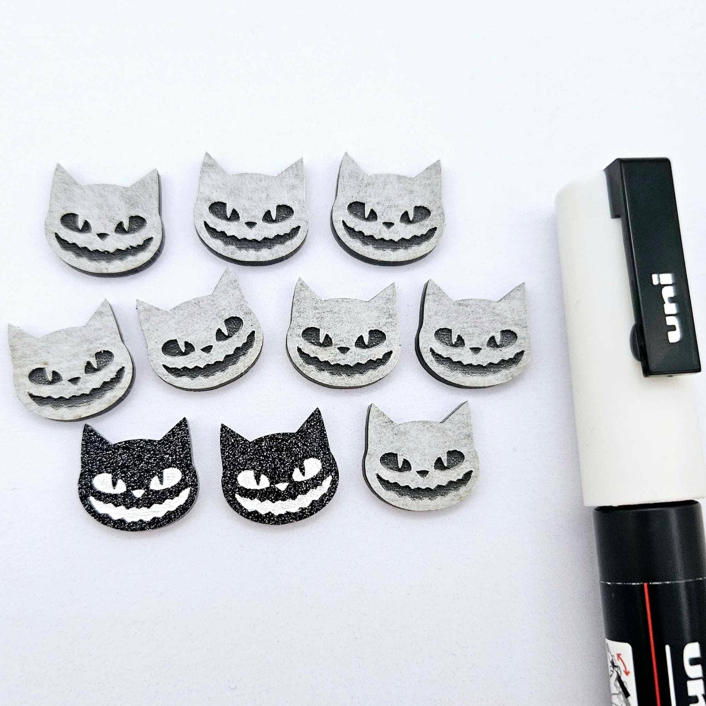 One pair of black glitter acrylic Spooky Cat stud earring blanks with white paint-filled details, surrounded by multiple unpainted pieces with protective paper on, pictured with a White Posca Paint Pen
