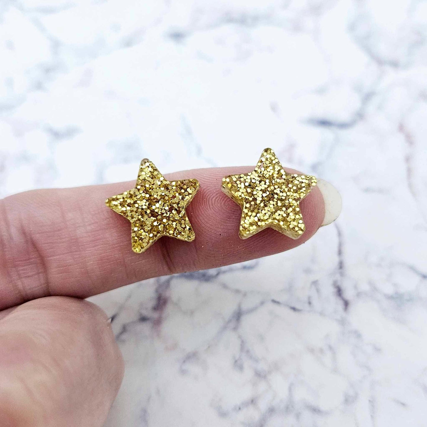One pair of 14mm gold glitter acrylic star stud earring blanks shown on a finger for size reference