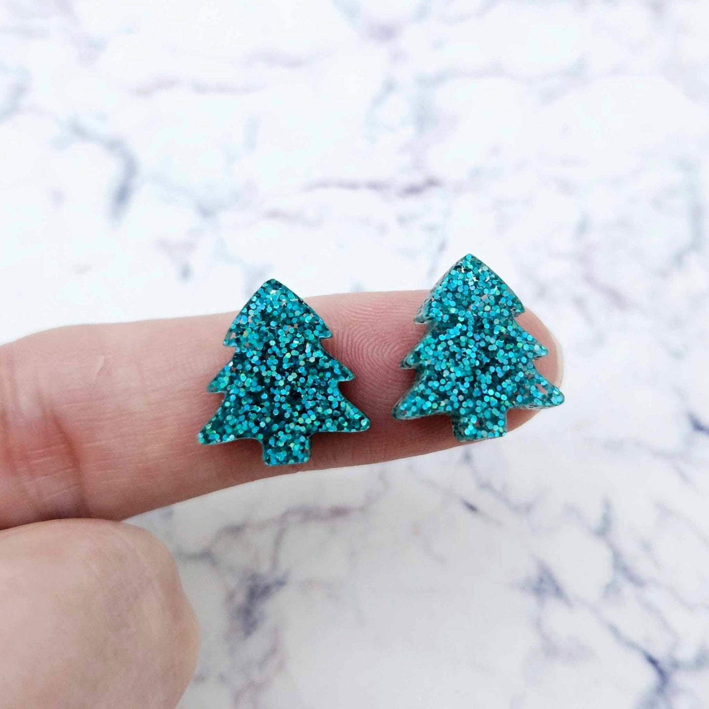 One pair of 16mm teal glitter Christmas tree stud earring blanks shown on a finger for size reference.