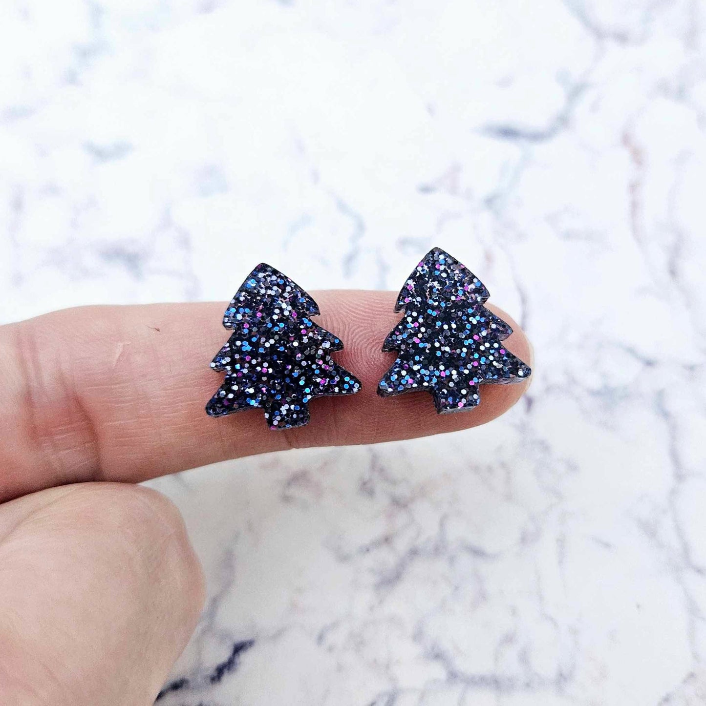 One pair of 16mm holographic blue glitter Christmas tree stud earring blanks shown on a finger for size reference.