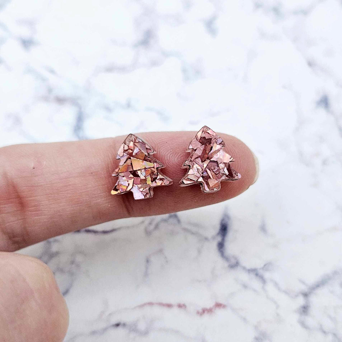 One pair of 12mm chunky holographic rose gold glitter acrylic Christmas tree stud earring blanks shown on a finger for size reference.