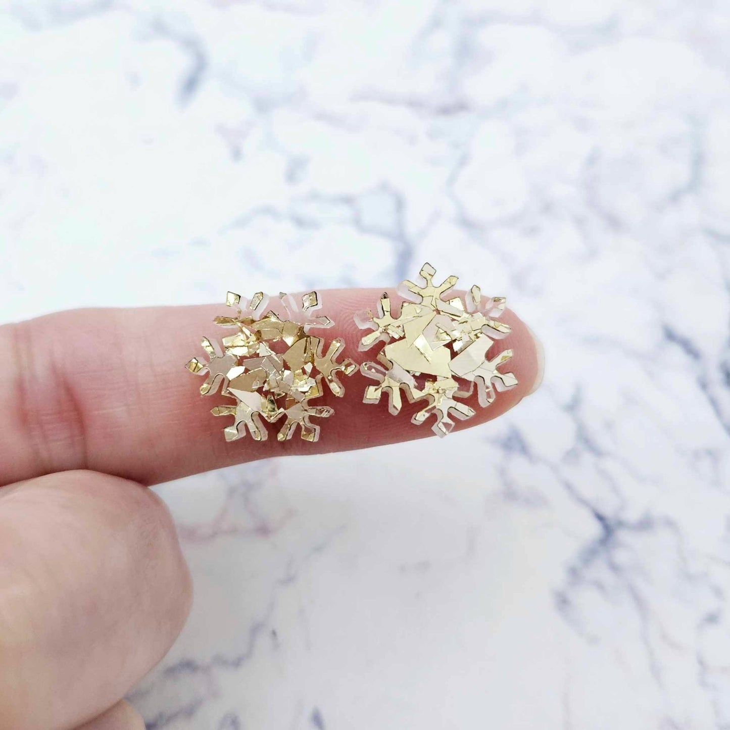 One pair of 16mm chunky gold glitter acrylic snowflake stud earring blanks shown on a finger for size reference.