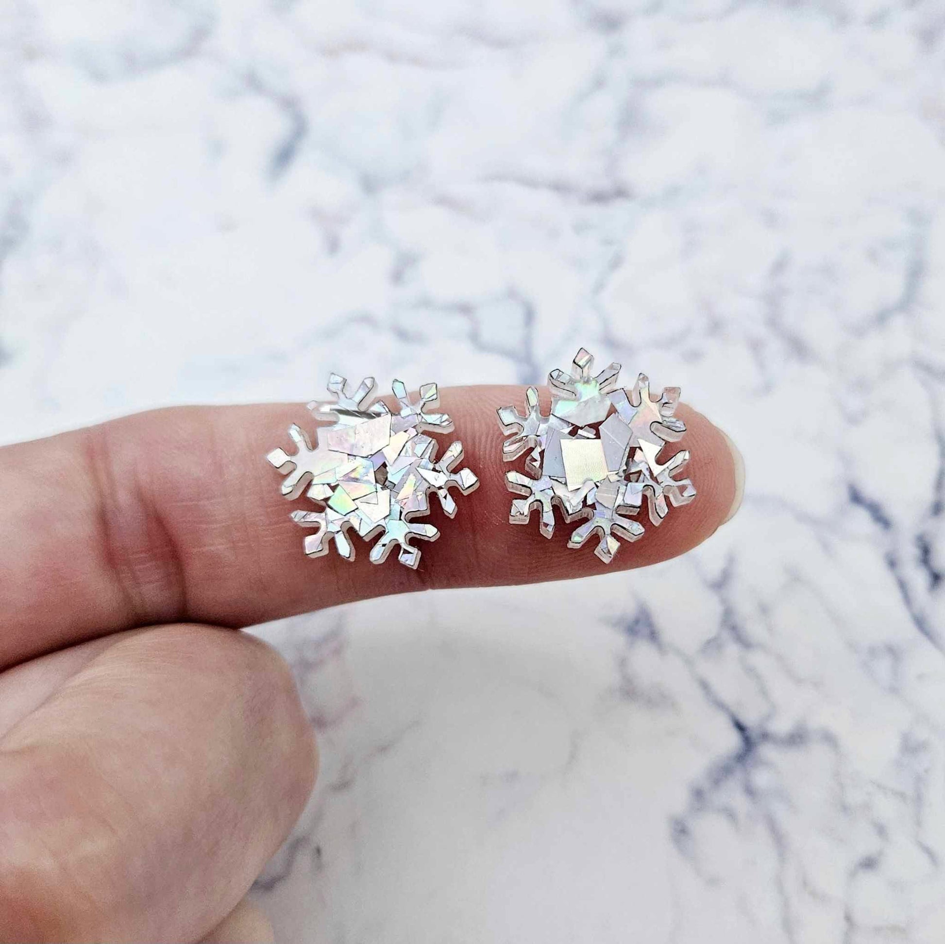 One pair of 16mm holographic silver glitter acrylic snowflake stud earring blanks shown on a finger for size reference.
