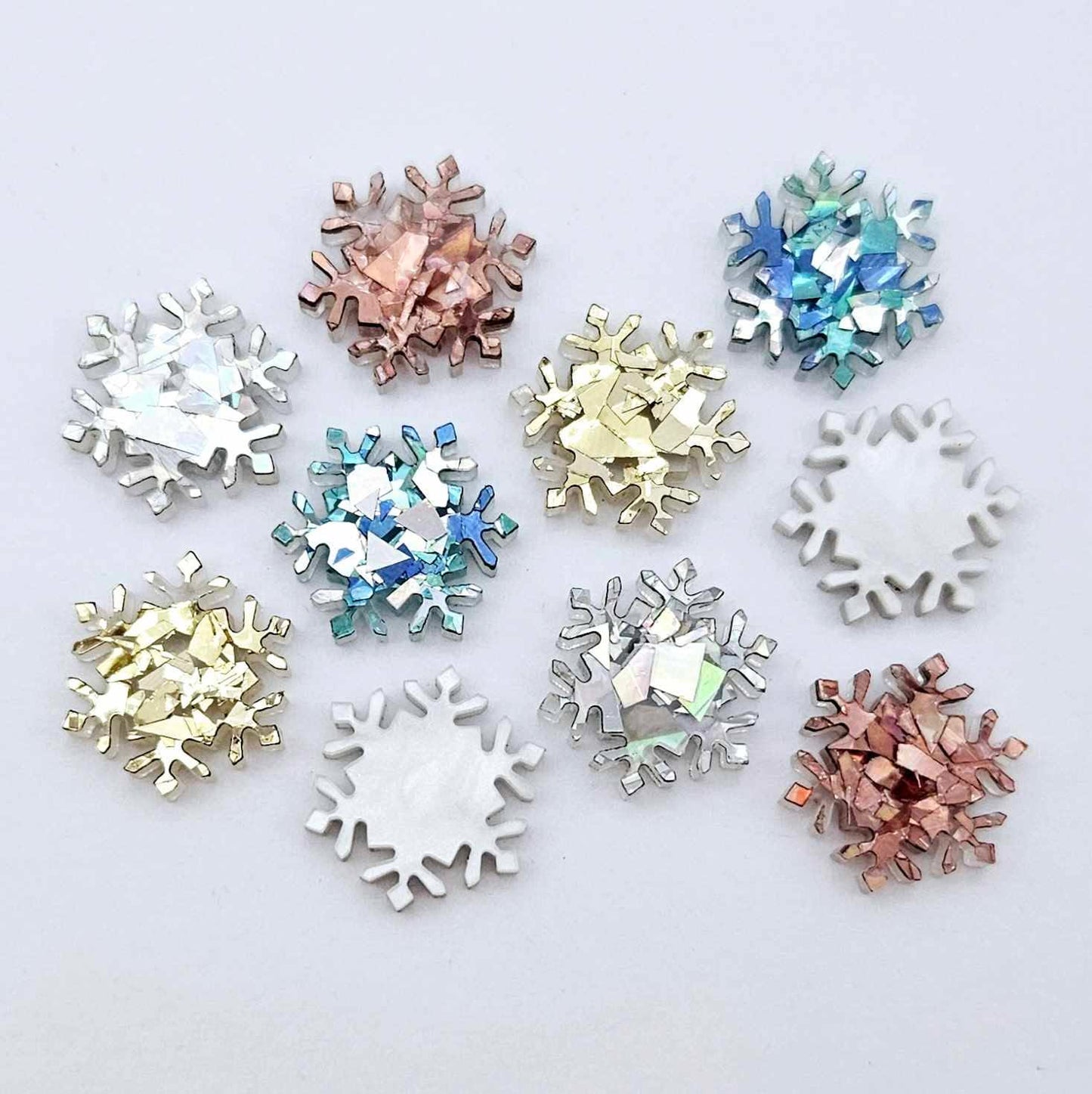 Five colours of 16mm snowflake stud earring blanks in holographic silver, aqua, chunky gold, holographic rose gold glitter and white marble acrylic laying flat.