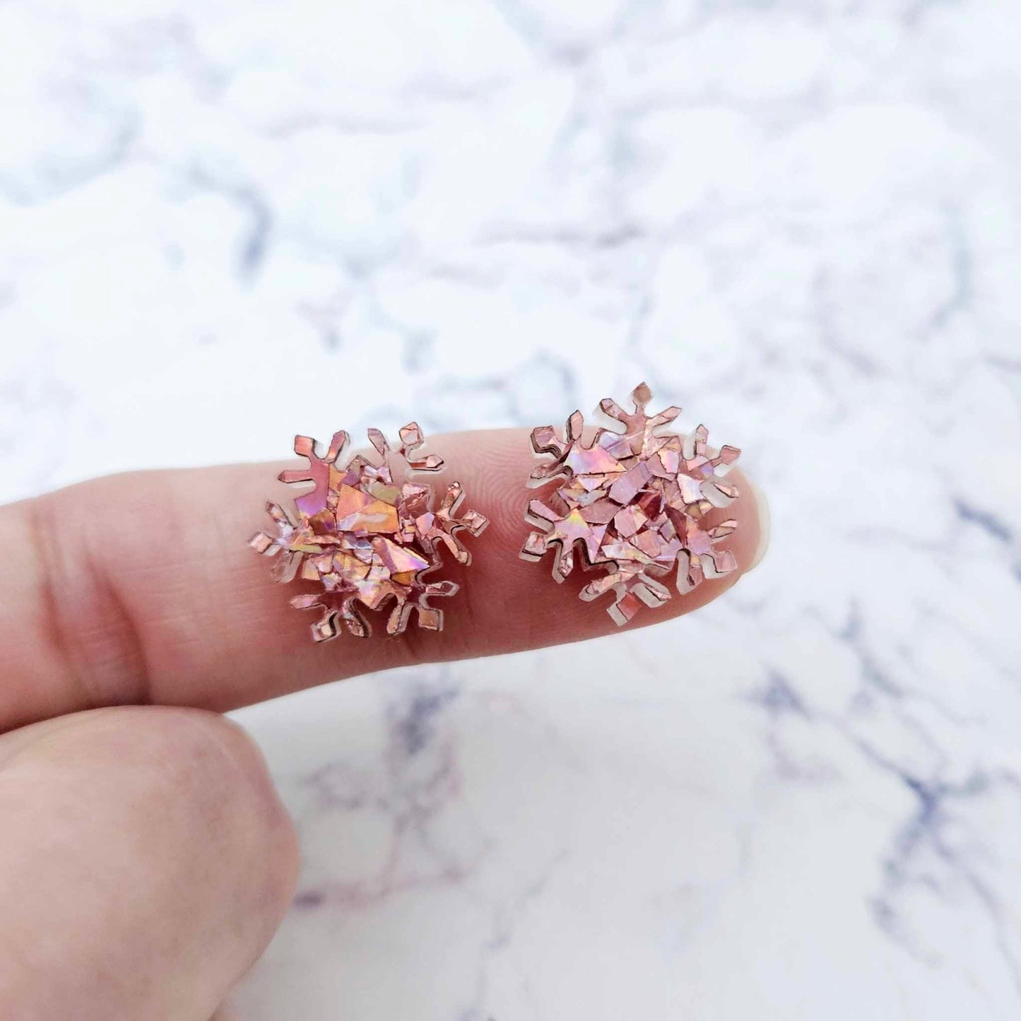 One pair of 16mm holographic rose gold glitter acrylic snowflake stud earring blanks shown on a finger for size reference.