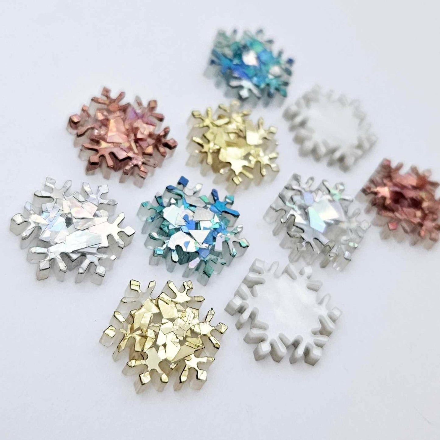 Five colours of 16mm snowflake stud earring blanks in holographic silver, aqua, chunky gold, holographic rose gold glitter and white marble acrylic laying flat.