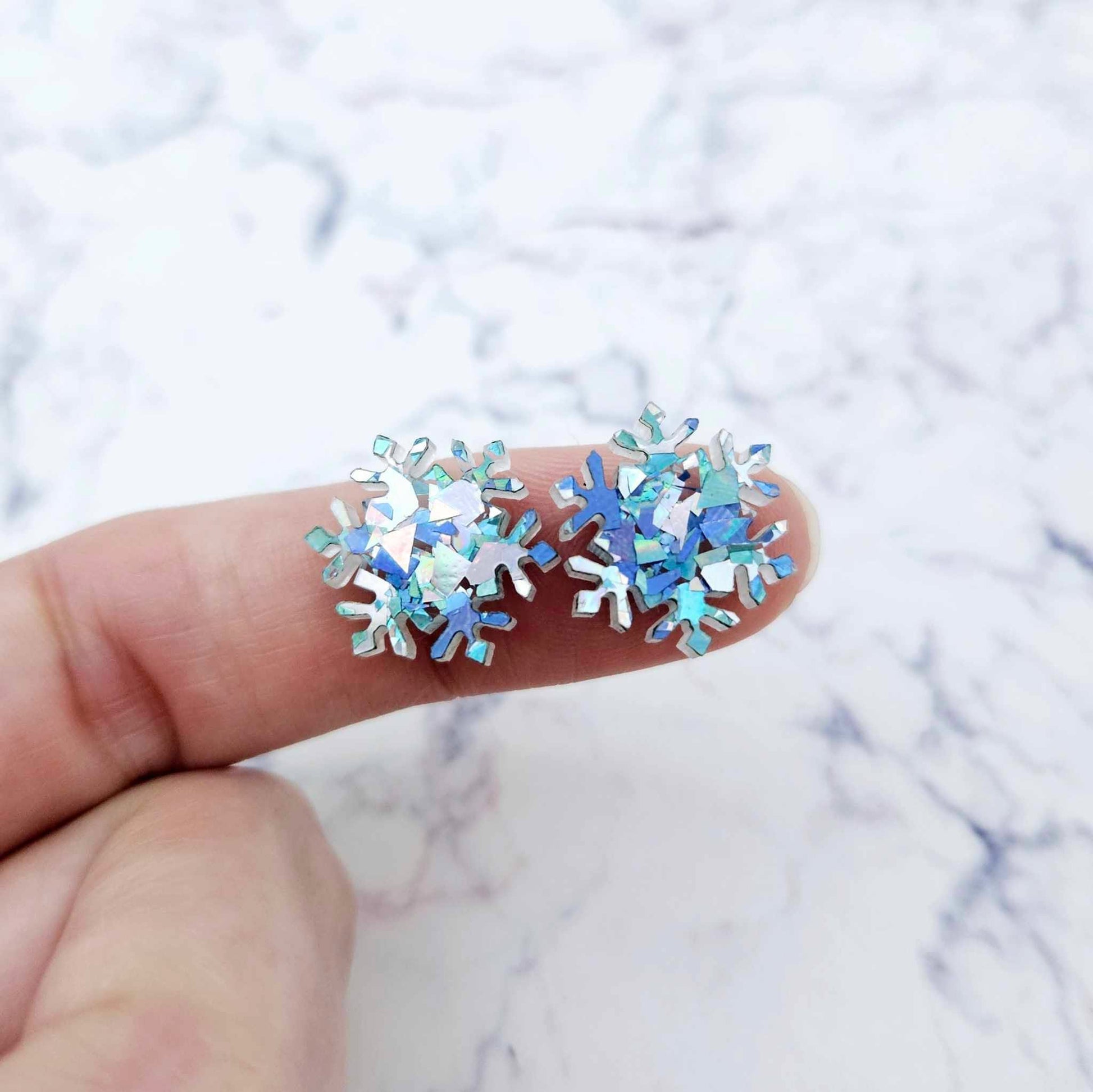 One pair of 16mm chunky aqua glitter acrylic snowflake stud earring blanks shown on a finger for size reference.