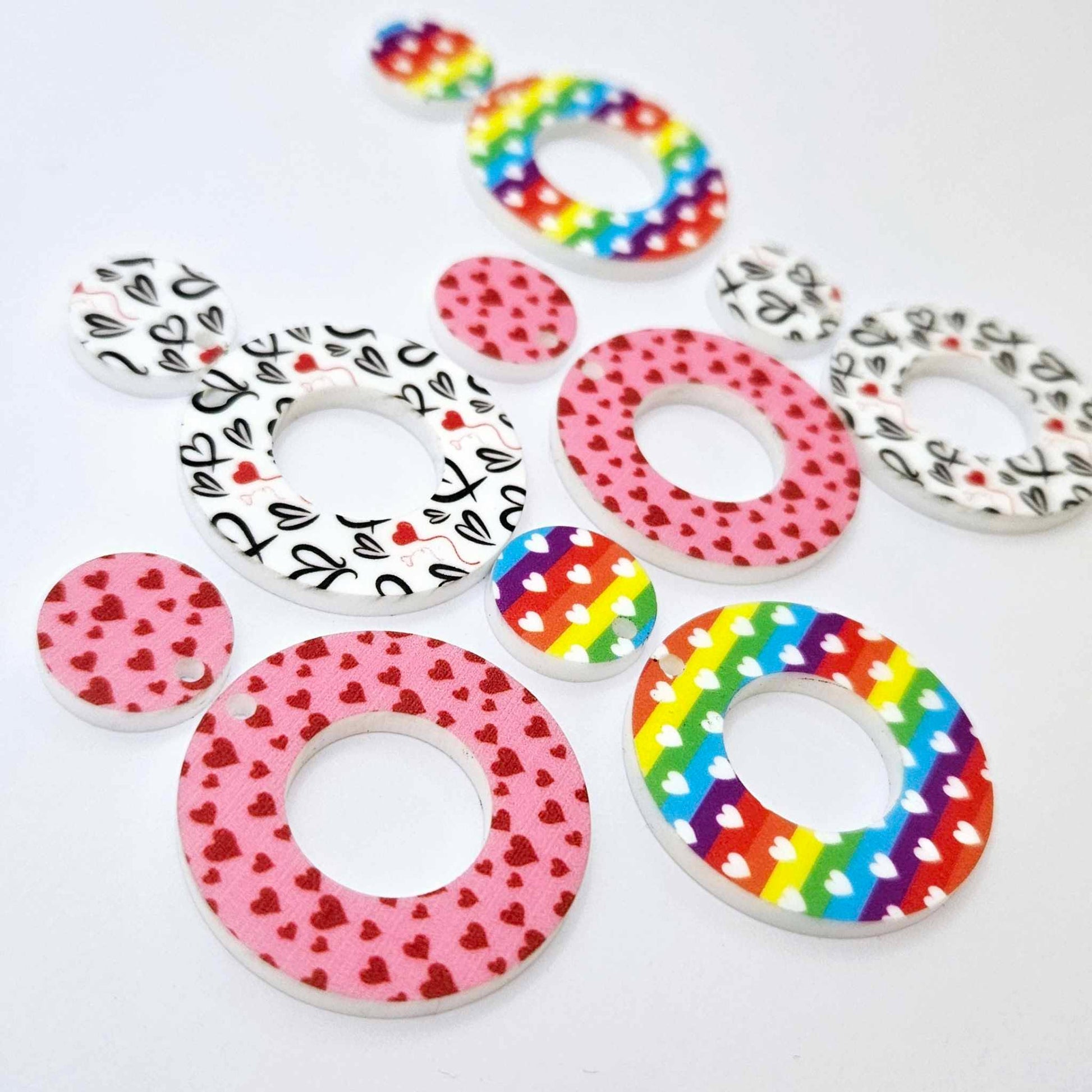 Three pairs of 33mm Love is Love UV printed acrylic mini hoop dangle earring blanks with matching toppers in Rainbow Love, Pink & Red Hearts and Red & Black Hearts designs laying flat.