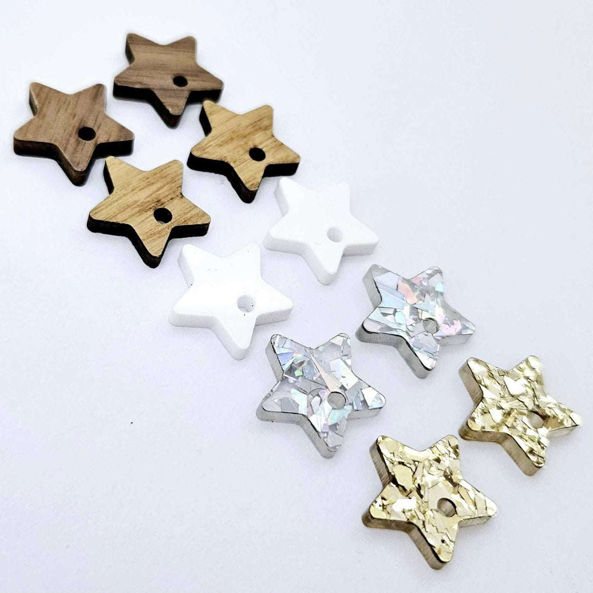 16mm star earring toppers in chunky gold glitter, holographic silver glitter, white acrylic, oak and walnut, with one pair of each finish laid flat together.