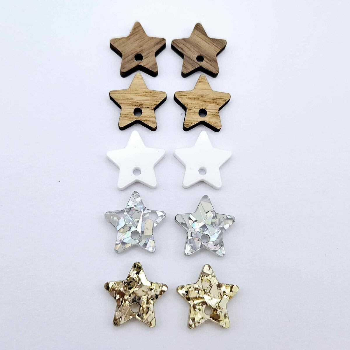 16mm star earring toppers in chunky gold glitter, holographic silver glitter, white acrylic, oak and walnut, with one pair of each finish laid flat together.