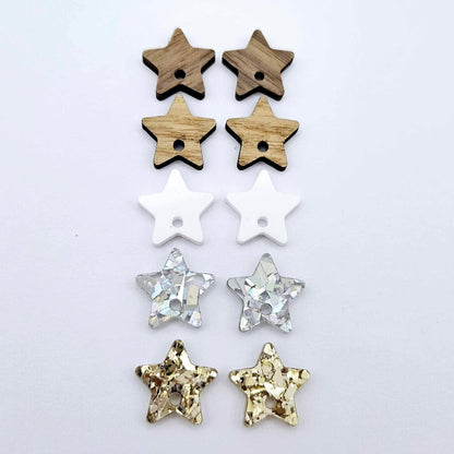16mm star earring toppers in chunky gold glitter, holographic silver glitter, white acrylic, oak and walnut, with one pair of each finish laid flat together.