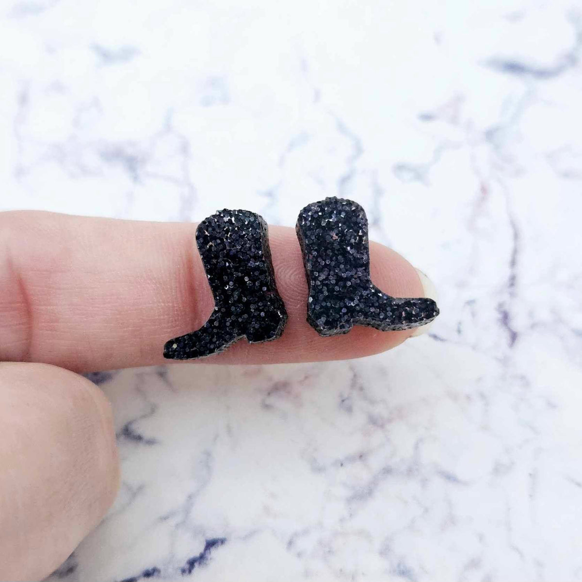 One pair of 15mm holographic black glitter acrylic boot stud earring blanks shown on a finger to for size reference.