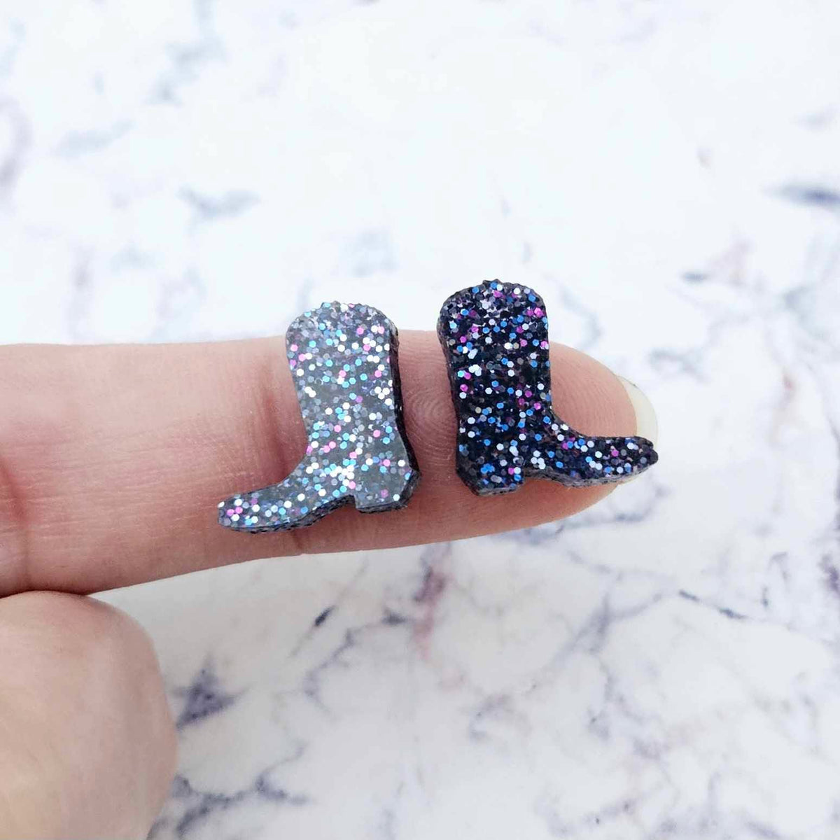 One pair of 15mm holographic blue glitter acrylic boot stud earring blanks shown on a finger to for size reference.