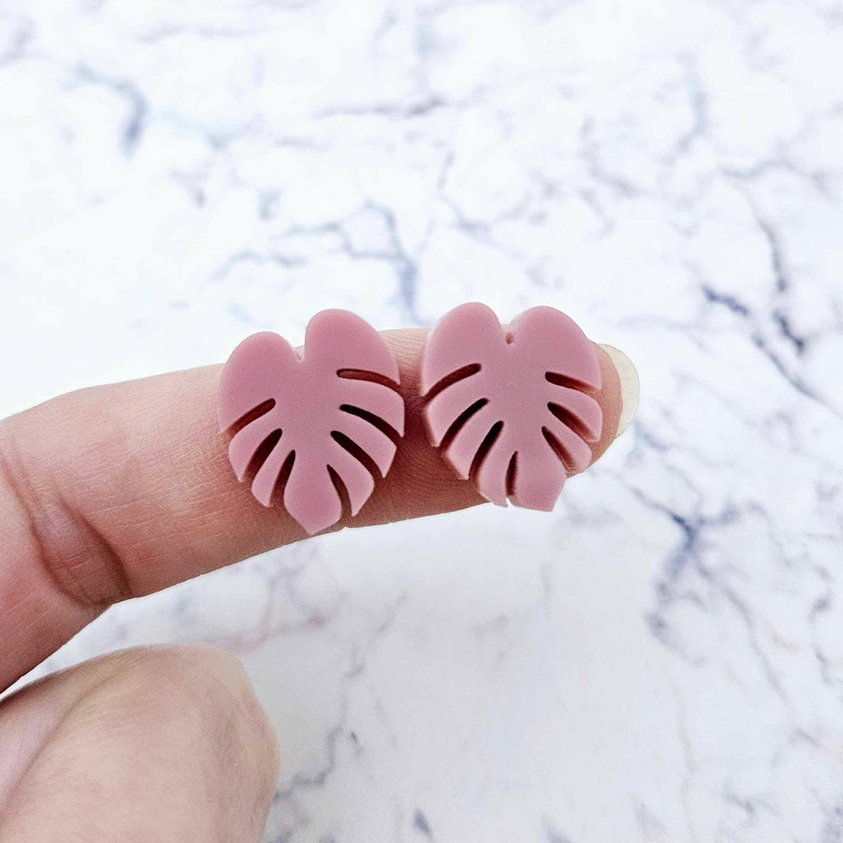 One pair of 17mm dusty rose pink acrylic monstera leaf stud earring blanks shown on a finger for size reference.