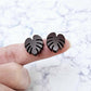 One pair of 17mm chocolate brown acrylic monstera leaf stud earring blanks shown on a finger for size reference.