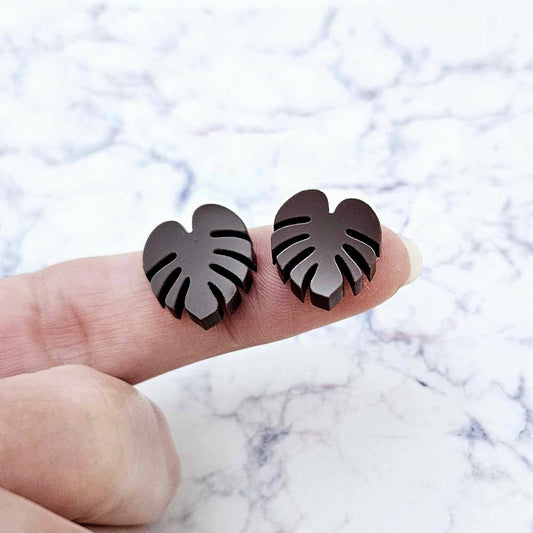 One pair of 17mm chocolate brown acrylic monstera leaf stud earring blanks shown on a finger for size reference.
