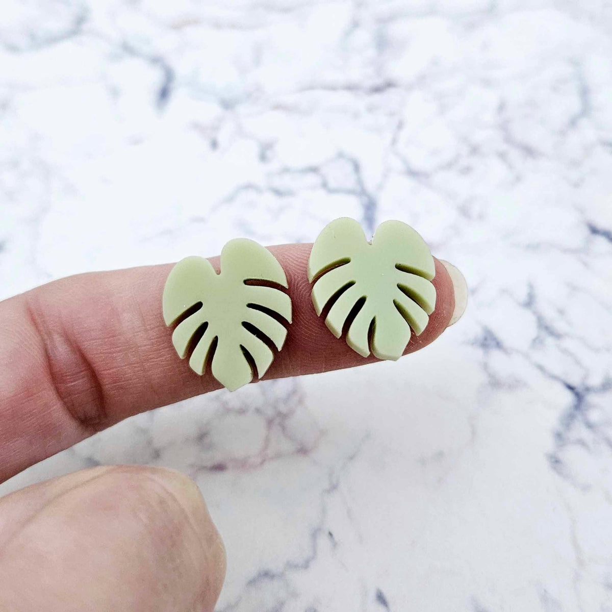 One pair of 17mm eucalyptus green acrylic monstera leaf stud earring blanks shown on a finger for size reference.