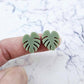 One pair of 17mm olive green acrylic monstera leaf stud earring blanks shown on a finger for size reference.
