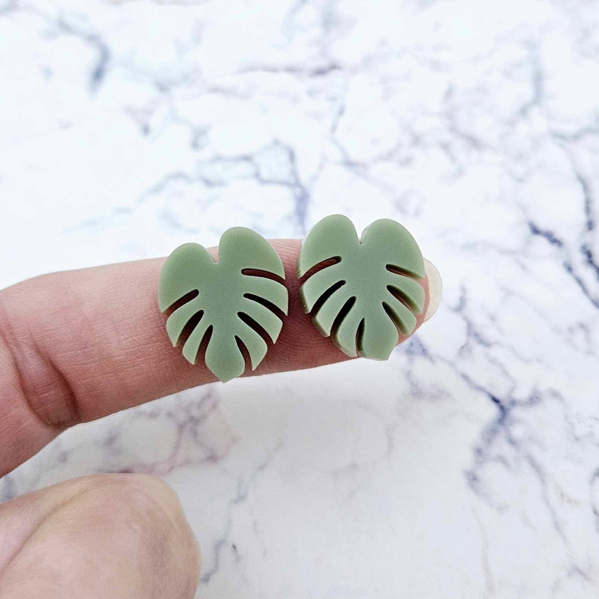 One pair of 17mm olive green acrylic monstera leaf stud earring blanks shown on a finger for size reference.