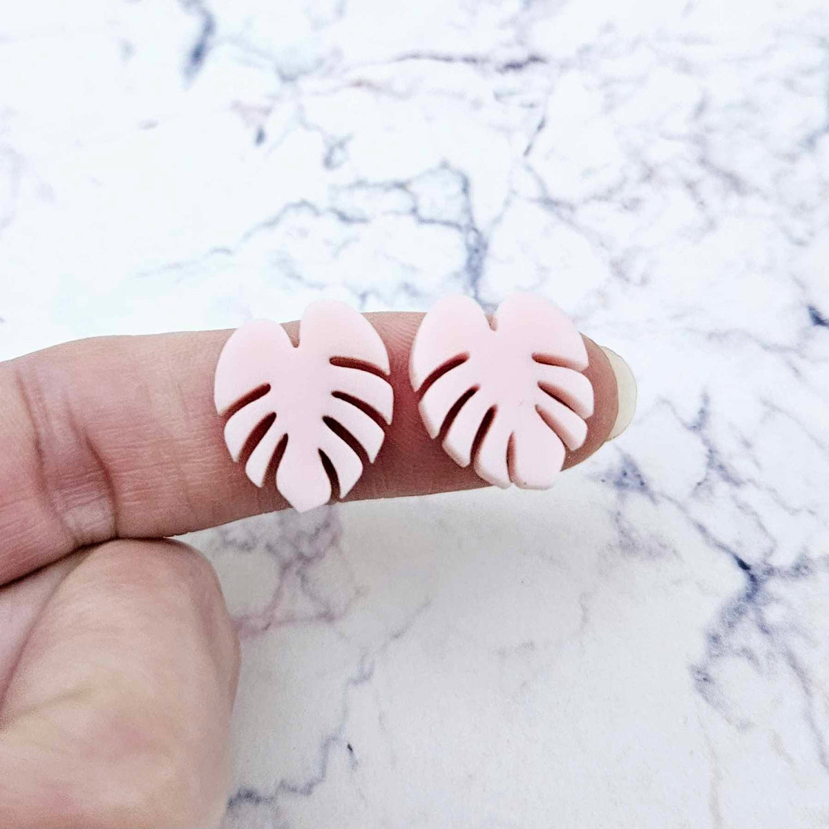 One pair of 17mm strawberry milkshake pink acrylic monstera leaf stud earring blanks shown on a finger for size reference.