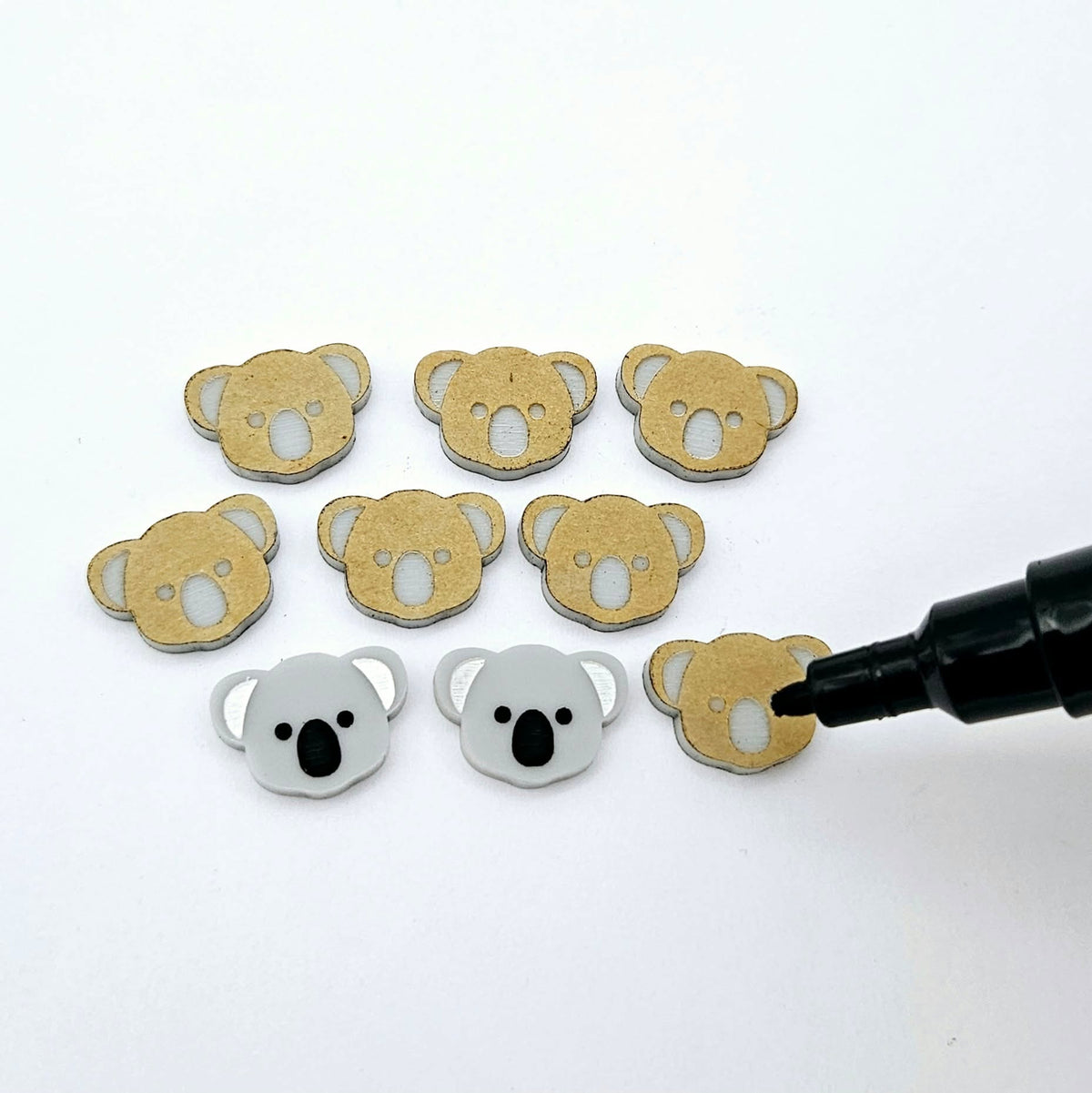 Solid Grey acrylic koala stud blanks showing one finished pair with protective paper removed and engraved details paint filled in black and white, alongside unpeeled blanks with protective paper still attached and a POSCA paint pen used for decorating.