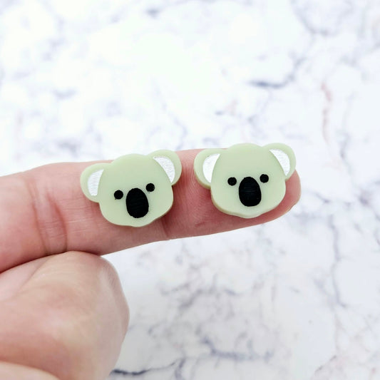 One pair of 18mm solid eucalyptus green acrylic koala stud blanks displayed on a finger to show their petite size, featuring engraved eyes, nose and ears with black and white paint-filled details.