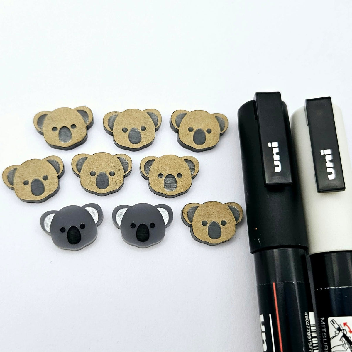 Solid charcoal grey acrylic koala stud blanks showing one finished pair with protective paper removed and engraved details paint filled in black and white, alongside unpeeled blanks with protective paper still attached and POSCA paint pens used for decorating.