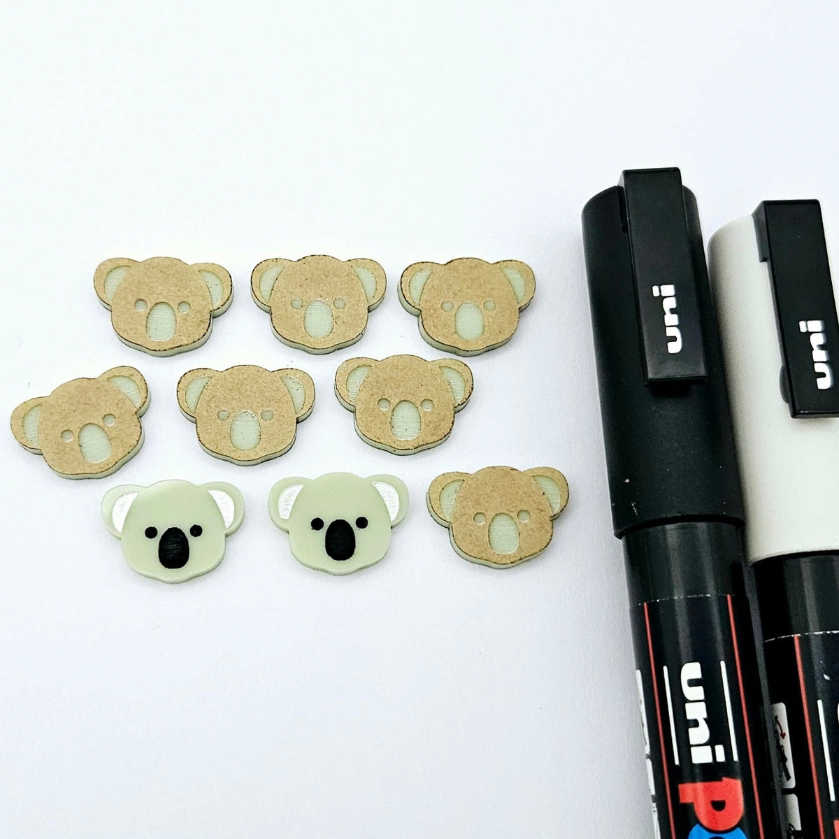 Solid Eucalyptus Green acrylic koala stud blanks showing one finished pair with protective paper removed and engraved details paint filled in black and white, alongside unpeeled blanks with protective paper still attached and POSCA paint pens used for decorating.