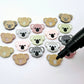 18mm solid acrylic koala stud earring blanks in Grey, Strawberry Milkshake, Dark Grey, Eucalyptus Earth and Dusty Rose, with five paint-filled pairs laid flat and additional unpainted koalas scattered around and an uncapped black Posca Pen.